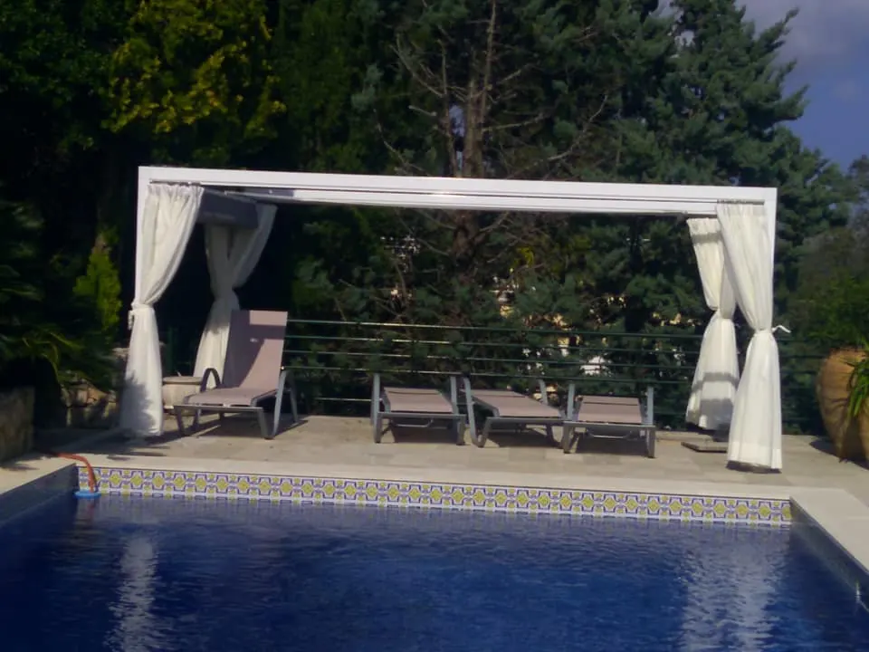 Pérgola blanca con cortinas junto a piscina con hamacas